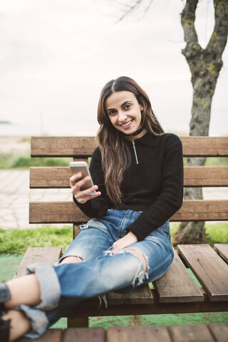 Porträt einer lächelnden jungen Frau mit Mobiltelefon auf einer Bank im Freien sitzend, lizenzfreies Stockfoto