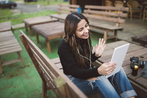 Lächelnde junge Frau sitzt auf einer Bank im Freien und benutzt ein Tablet und Kopfhörer für einen Video-Chat, lizenzfreies Stockfoto