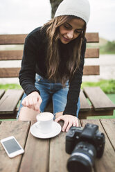 Junge Fotografin sitzt auf einer Bank im Freien mit einer Tasse Kaffee - RAEF02214