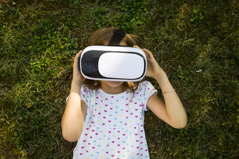 Kleines Mädchen mit Virtual-Reality-Brille auf der Wiese im Garten liegend, lizenzfreies Stockfoto