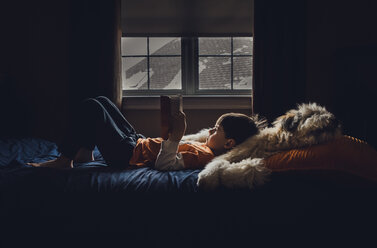 Seitenansicht eines Jungen, der ein Buch liest, während er zu Hause auf dem Bett liegt - CAVF51056