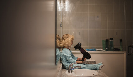 Seitenansicht eines Mädchens, das einen Haartrockner hält, während es zu Hause am Waschbecken sitzt - CAVF51140