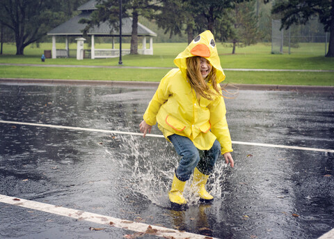 Verspieltes Mädchen trägt Regenmantel und springt bei Regen in eine Pfütze, lizenzfreies Stockfoto