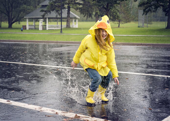 Verspieltes Mädchen trägt Regenmantel und springt bei Regen in eine Pfütze - CAVF51142