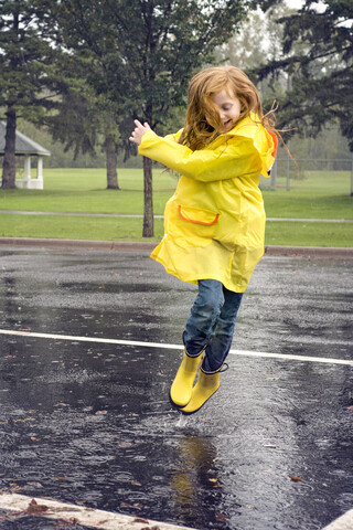 Fröhliches Mädchen mit Regenmantel, das bei Regen auf die Straße springt, lizenzfreies Stockfoto