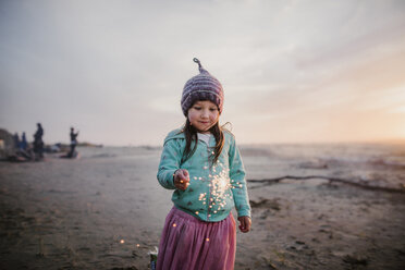 Girl wearing knit hat while holding illuminated sparkler against sky at beach - CAVF51176