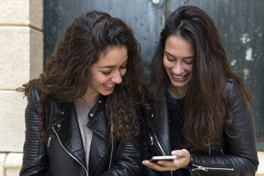 Glückliche Freunde, die ein Mobiltelefon benutzen, während sie vor der Tür sitzen - CAVF51211