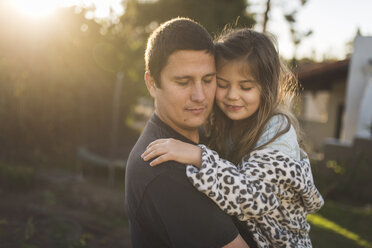Vater und Tochter mit geschlossenen Augen umarmen sich im Garten bei Sonnenuntergang - CAVF51221