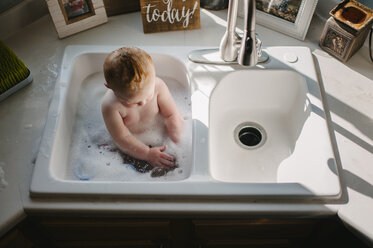 High angle view of shirtless baby boy sitting in kitchen sink at home - CAVF51247