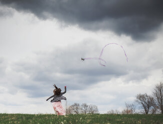 Rückansicht eines verspielten Mädchens, das einen Drachen gegen einen bewölkten Himmel im Park steigen lässt - CAVF51336