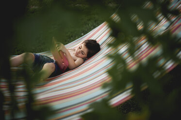 High angle view of boy looking at slate while lying on hammock at backyard seen through branches - CAVF51338