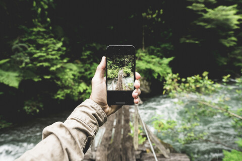 Ausgeschnittene Hand eines Wanderers, der mit seinem Smartphone einen Steg über einen Fluss im Wald fotografiert, lizenzfreies Stockfoto