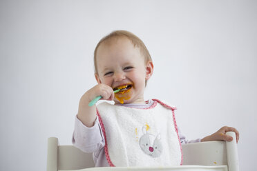 Portrait of laughing baby girl on high chair eating mush - JLOF00285