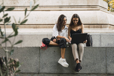 Zwei Freunde sitzen Seite an Seite an einer Wand und benutzen einen Laptop - KKAF02829