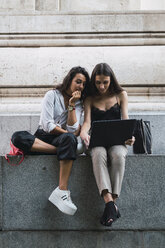 Zwei Freunde sitzen Seite an Seite an einer Wand und benutzen einen Laptop - KKAF02830