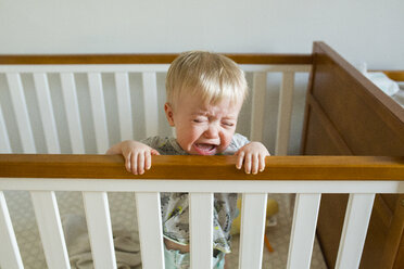 Baby boy crying while standing in crib at home - CAVF51365