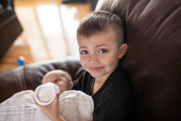 Porträt eines Jungen, der seinem Bruder Milch aus einer Babyflasche gibt, während er zu Hause auf dem Sofa sitzt - CAVF51376