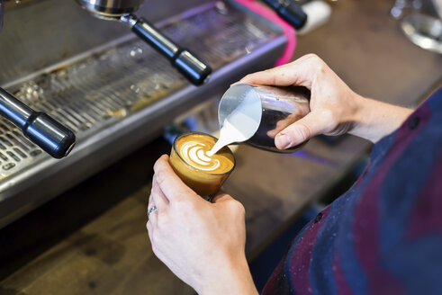 Mittelteil eines Barista, der Milch in den Kaffee gießt, während er ein schaumiges Getränk im Café zubereitet - CAVF51409