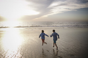 Rear view of playful brothers running at beach against sky during sunset - CAVF51412