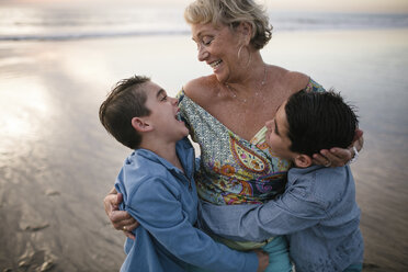 Happy grandmother with grandsons standing at beach during sunset - CAVF51413