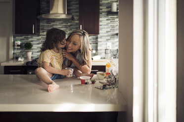 Girl kissing mother sitting on kitchen counter at home - CAVF51423