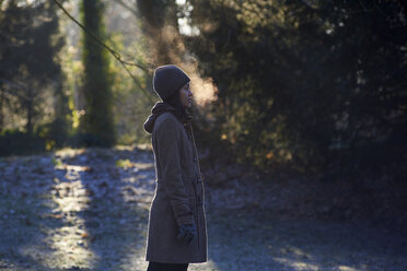 Side view of woman exhaling breath vapor while standing at park during winter - CAVF51443