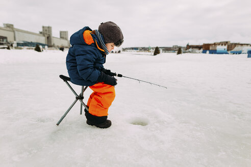 Side view of boy fishing in frozen lake while sitting on stool against sky - CAVF51454
