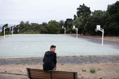 Männlicher jugendlicher Basketballspieler, der von einer Parkbank am Basketballplatz zurückblickt - CUF46442