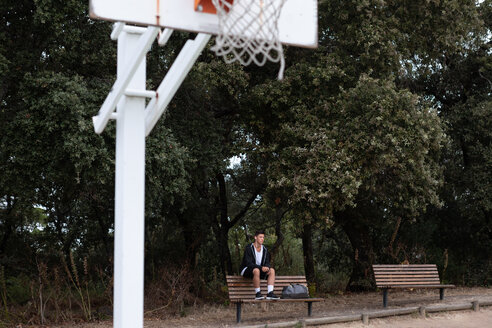 Männlicher jugendlicher Basketballspieler sitzt auf einer Parkbank am Basketballplatz - CUF46443