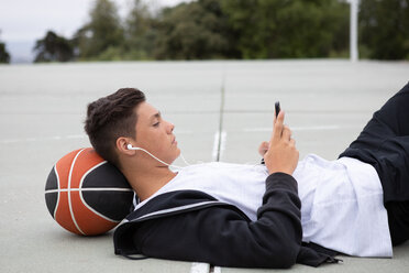 Männlicher Basketballspieler im Teenageralter, der auf dem Basketballplatz liegt und auf sein Smartphone schaut - CUF46446
