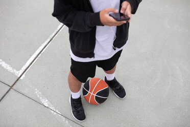 Männlicher jugendlicher Basketballspieler steht auf dem Basketballplatz und schaut auf sein Smartphone, Taille nach unten - CUF46447