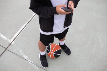 Male teenage basketball player standing on basketball court looking at smartphone, waist down - CUF46447