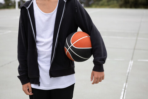 Männlicher Teenager-Basketballspieler auf dem Basketballplatz mit Ball unter dem Arm, Mittelteil, lizenzfreies Stockfoto