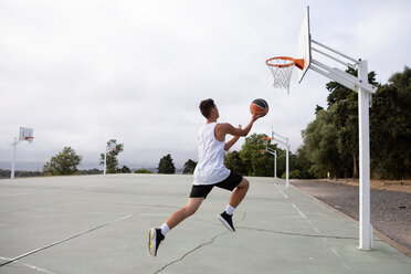 Male teenage basketball player jumping with ball toward basketball hoop - CUF46452