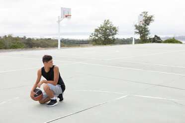 Männlicher jugendlicher Basketballspieler mit Ball auf dem Basketballplatz hockend - CUF46455