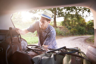 Young man in trilby removing luggage from car boot on rural road - CUF46563
