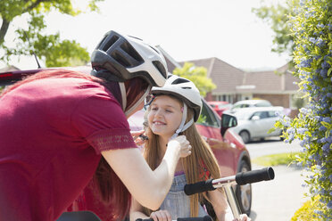 Mother fastening helmet on daughter riding scooter in sunny driveway - CAIF22193