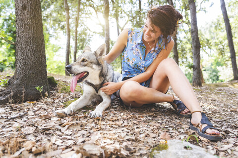 Lächelnde junge Frau sitzt auf dem Waldboden und streichelt ihren Hund, lizenzfreies Stockfoto