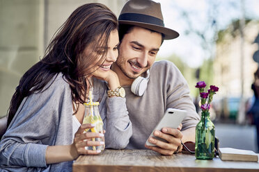 Glückliches junges Paar schaut in einem Café im Freien auf sein Handy - BSZF00786