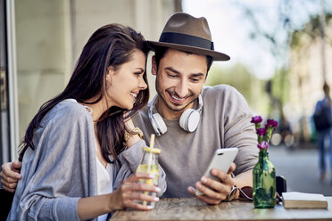 Glückliches junges Paar schaut in einem Café im Freien auf sein Handy - BSZF00787