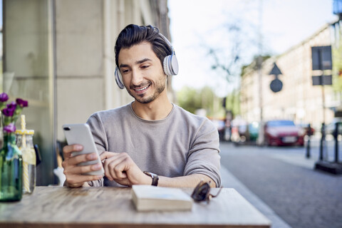 Lächelnder junger Mann, der in einem Café im Freien über Kopfhörer Musik hört, lizenzfreies Stockfoto