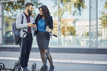 Smiling businessman and businesswoman with scooters talking on pavement - ZEDF01700