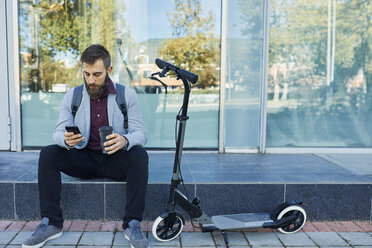 Businessman sitting outdoors using cell phone next to scooter - ZEDF01702