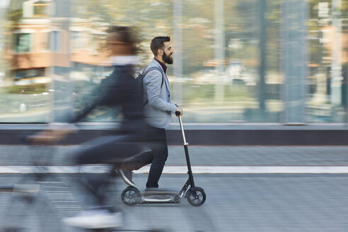 Businessman riding scooter along office building - ZEDF01712