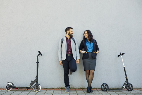 Smiling businessman and businesswoman with scooters leaning against a wall stock photo
