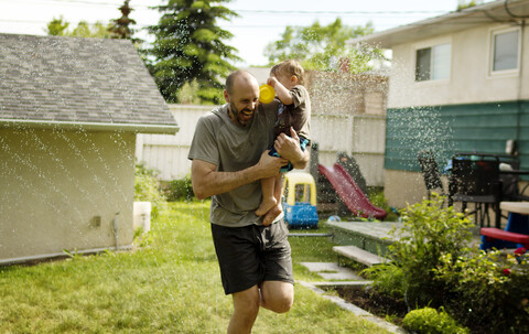 Fröhlicher Vater trägt seinen Sohn beim Spielen im Sprinkler im Garten, lizenzfreies Stockfoto