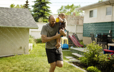 Fröhlicher Vater trägt seinen Sohn beim Spielen im Sprinkler im Garten - CAVF51463