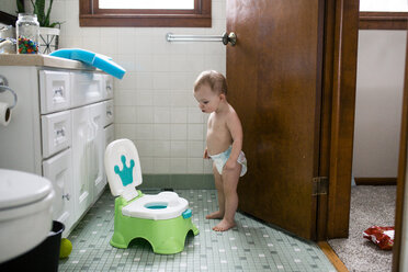 Shirtless baby girl looking at potty while standing in bathroom - CAVF51481