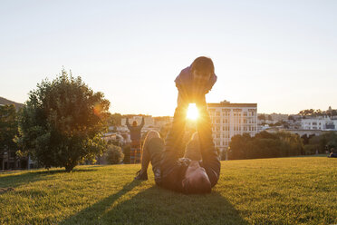 Playful father lifting daughter while lying on grassy field at park during sunny day - CAVF51494