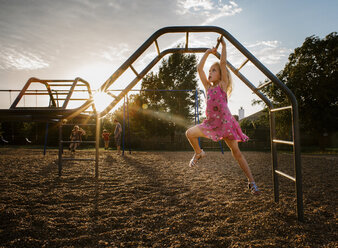 Mädchen, das auf dem Spielplatz bei Sonnenuntergang an einem Klettergerüst gegen den Himmel hängt - CAVF51511